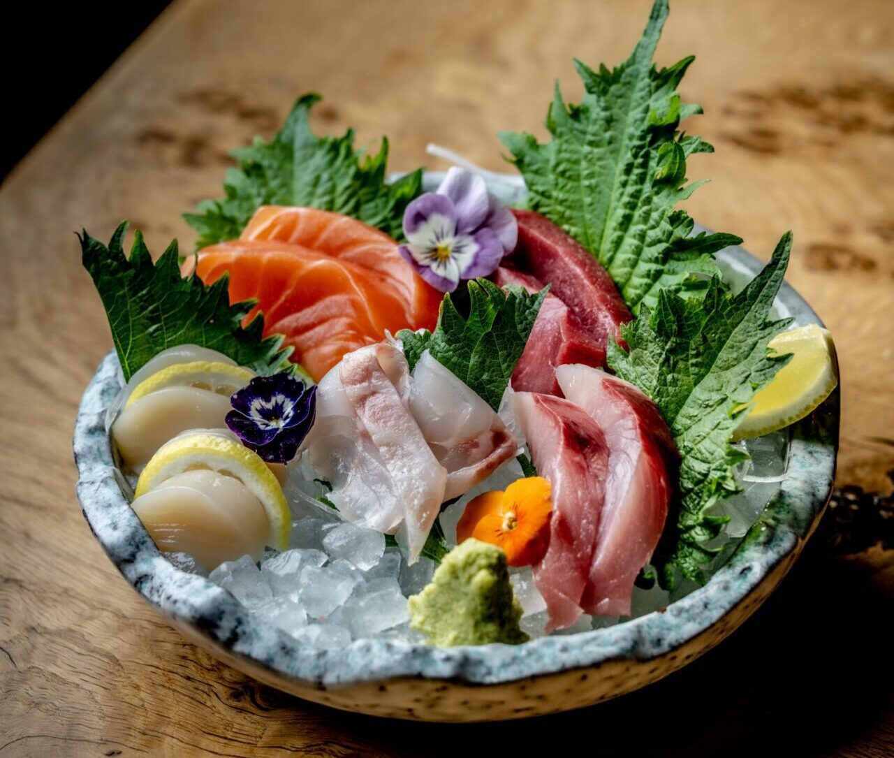 A plate of assorted sashimi, including salmon, tuna, and white fish, garnished with edible flowers, lemon wedges, and wasabi.