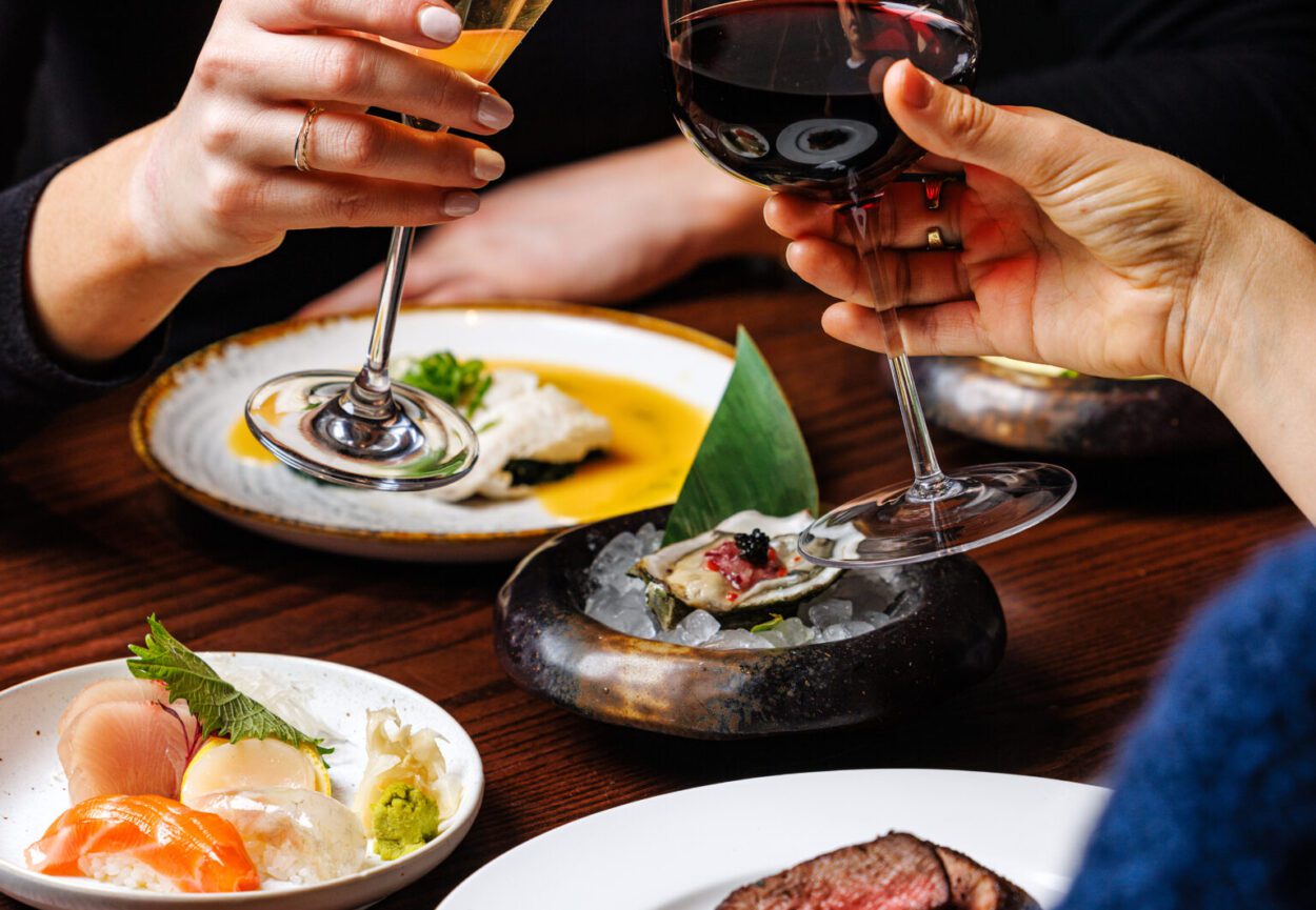 Two glasses of wine being held in a toasting position over four plates of Japanese cuisine.