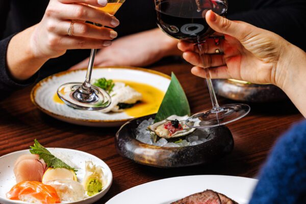 Two glasses of wine being held in a toasting position over four plates of Japanese cuisine.