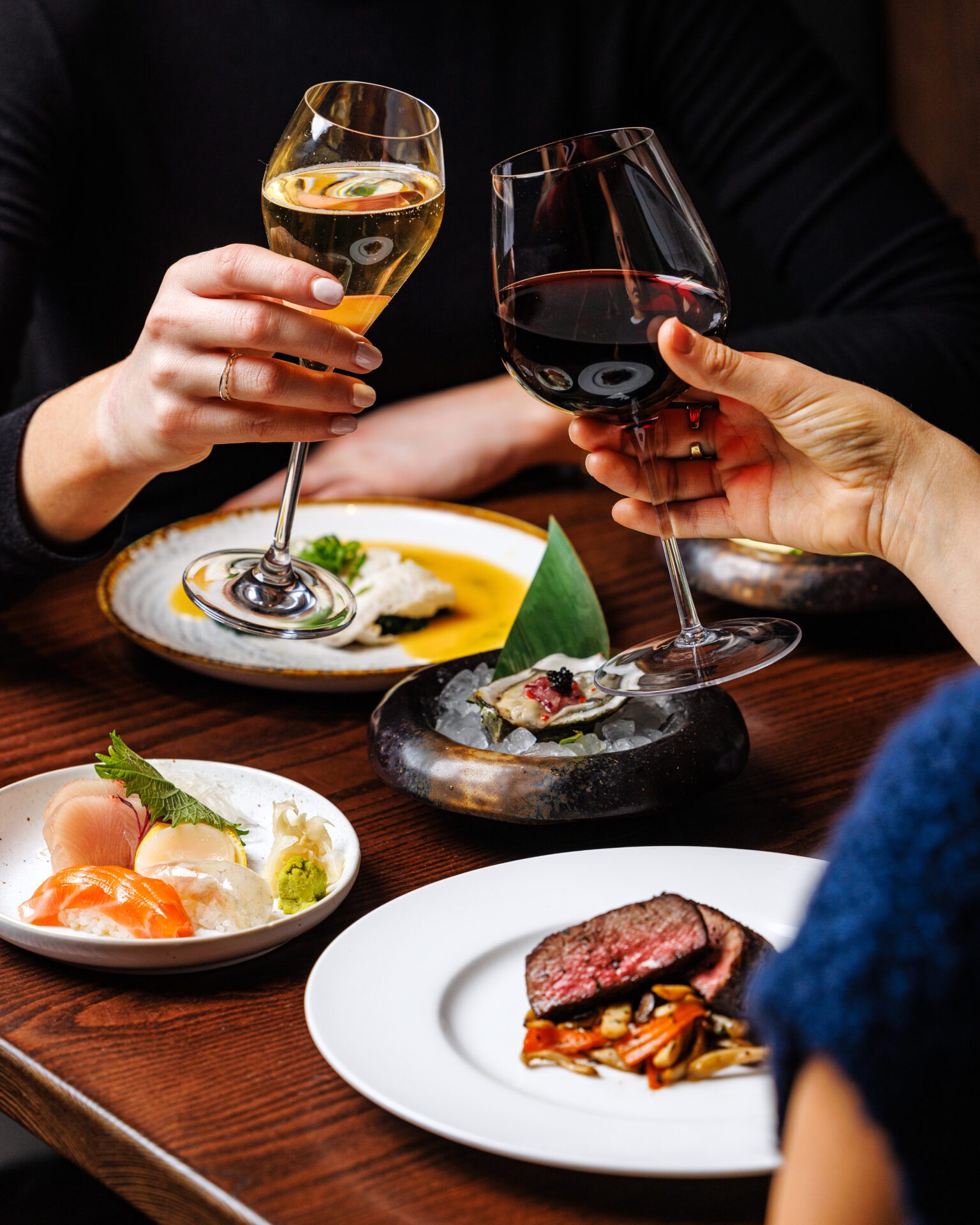 Two glasses of wine being held in a toasting position over four plates of Japanese cuisine.